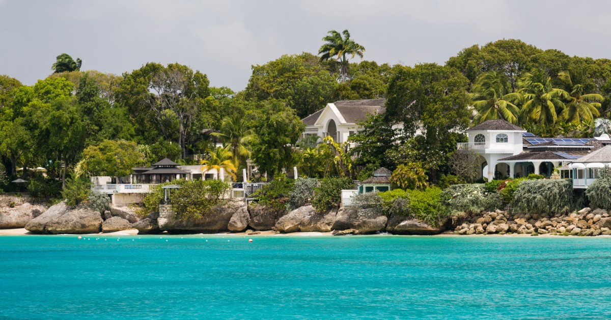 A row of houses that are off the coast of Barbados. There are multiple trees and rocks around the properties.