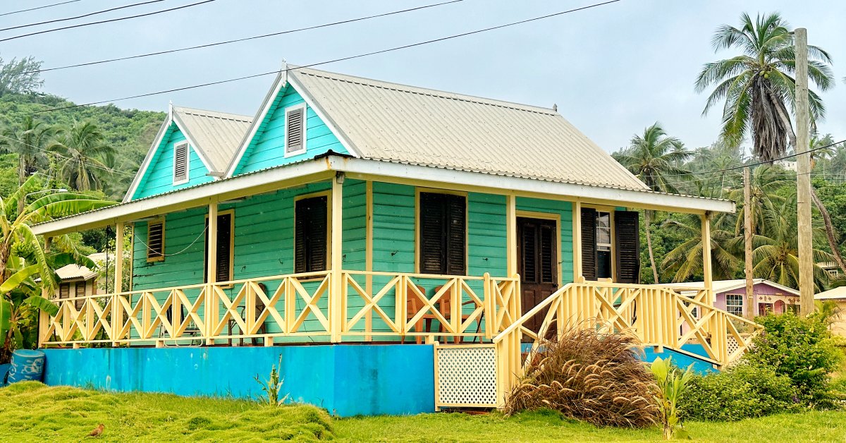 A blue house with a yellow railing. There are palm trees and bushes near the home, and the sky is cloudy.