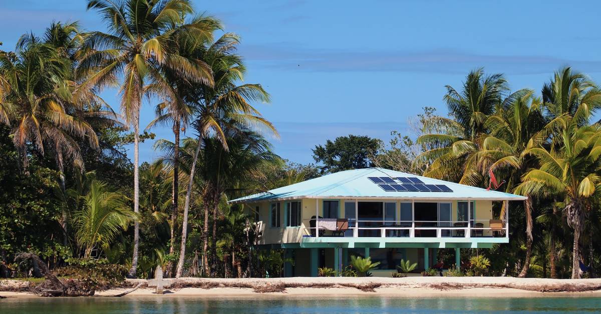 A two-story home on a beach with large, tall palm trees. The house is yellow and blue with solar panels on the roof.