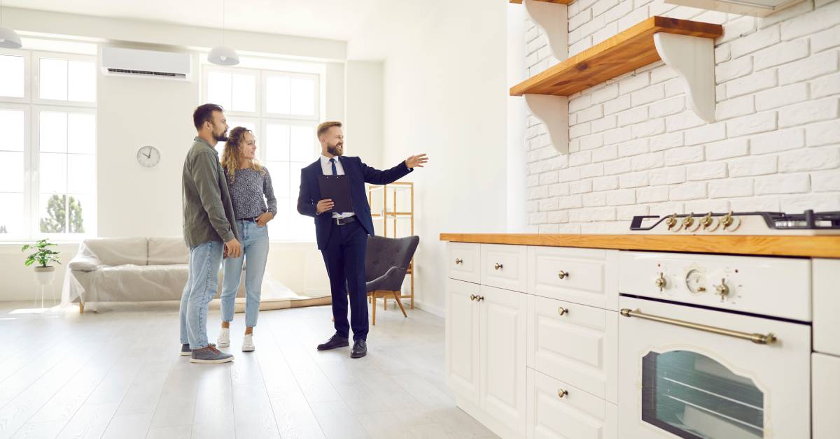 A man in a suit showing a home to a couple. The home's walls, ceiling, kitchen cabinets, and wooden floors are all white.