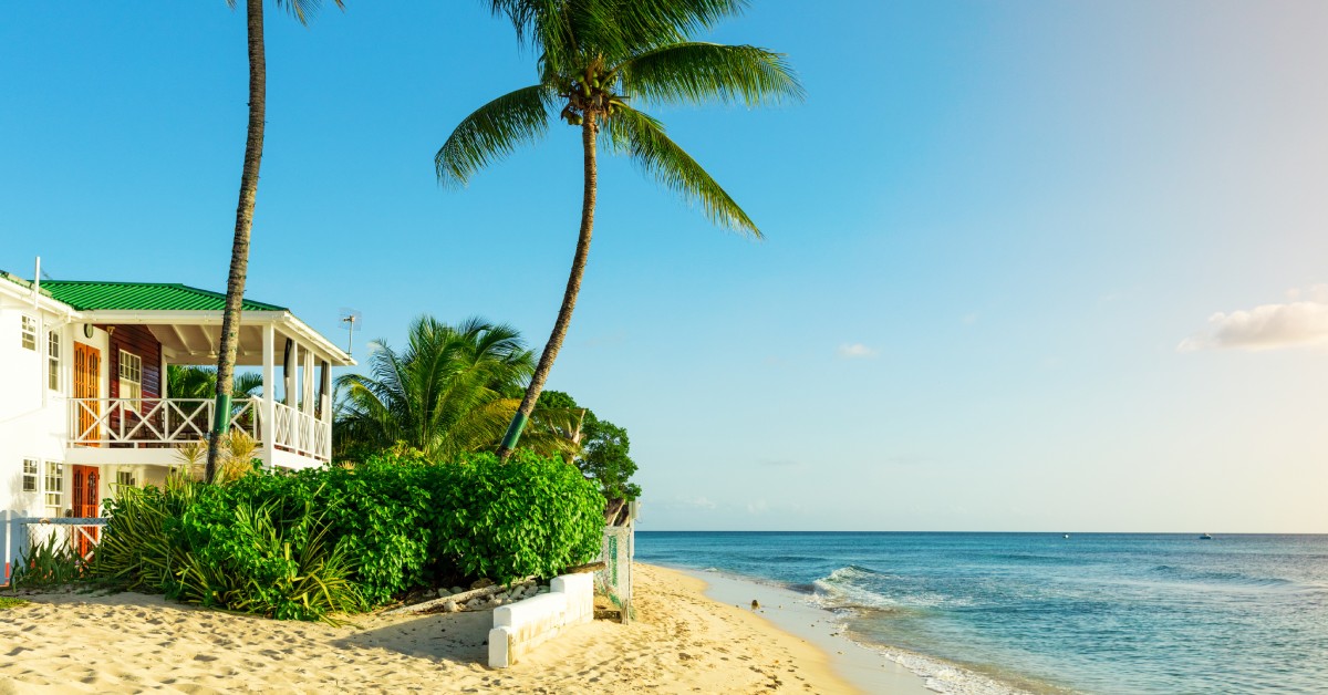 A quaint Barbados property on the edge of the beach with views of the sand, ocean, and tall palm trees.