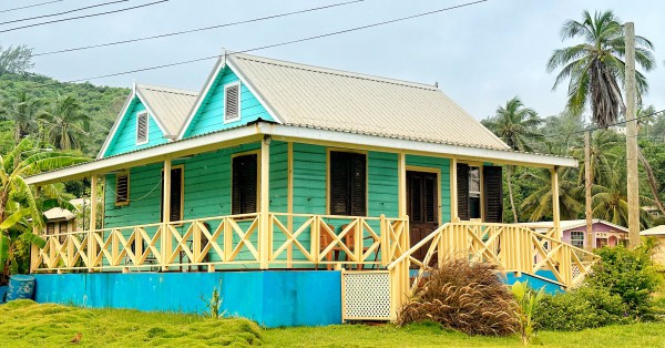 A blue house with a yellow railing. There are palm trees and bushes near the home, and the sky is cloudy.