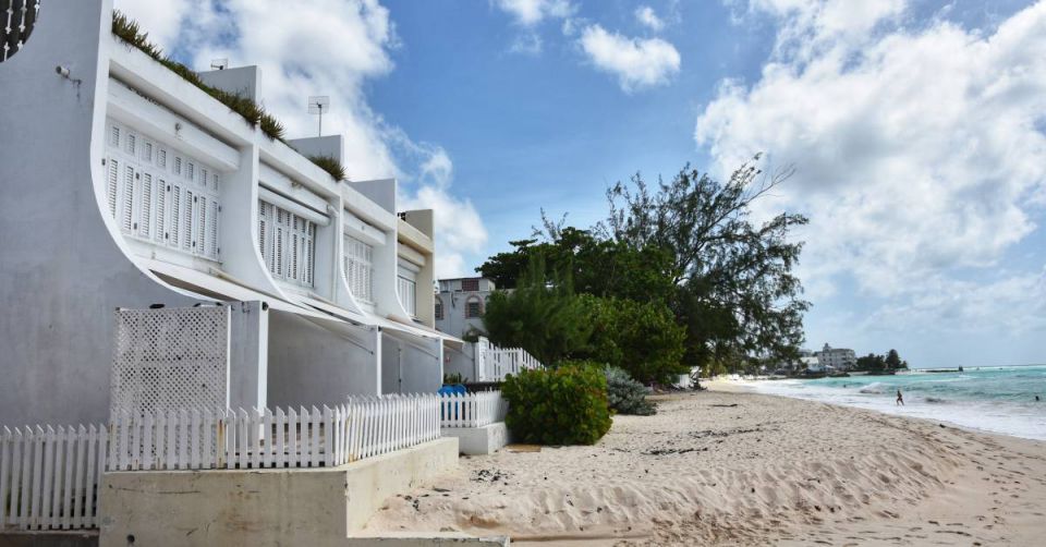 A white house by the beach. There's a fence around the house, and there are white clouds in the sky.