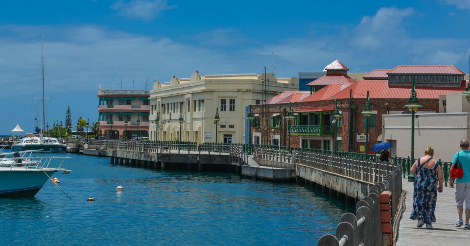 A man and a woman walking along a boardwalk near bright blue water. Colorful buildings line the boardwalk.